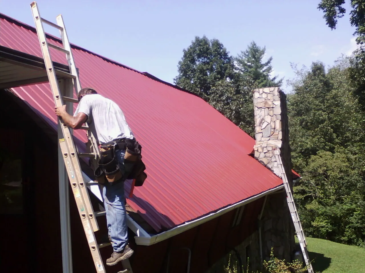Roofer on a ladder beside a red metal roof and stone chimney — Soffit & Fascia Repair by Roofing Brothers in Braidwood